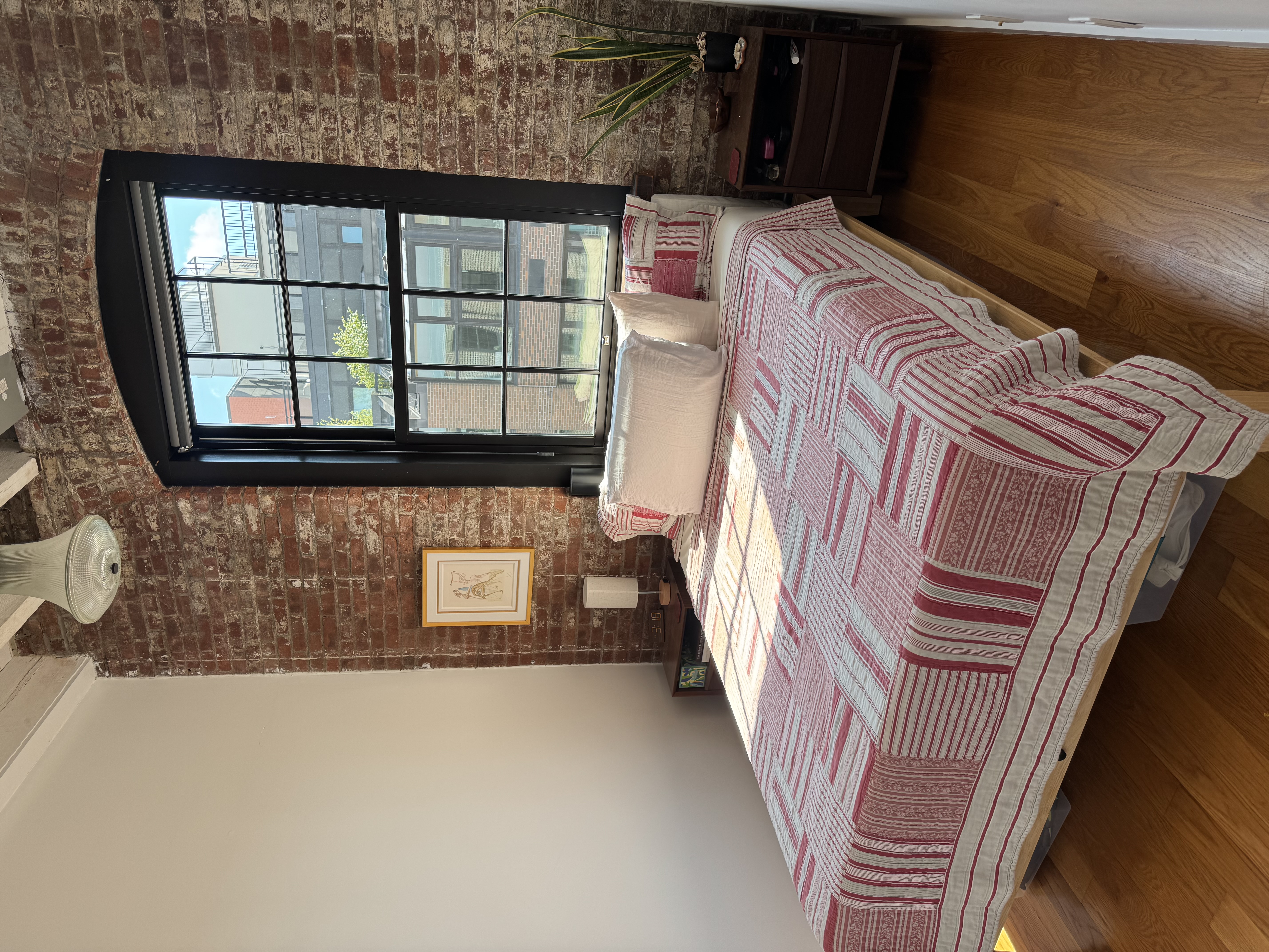 Main bedroom with a queen bed in a red and white patchwork quilt against exposed brick, sunlight coming through the paned window
