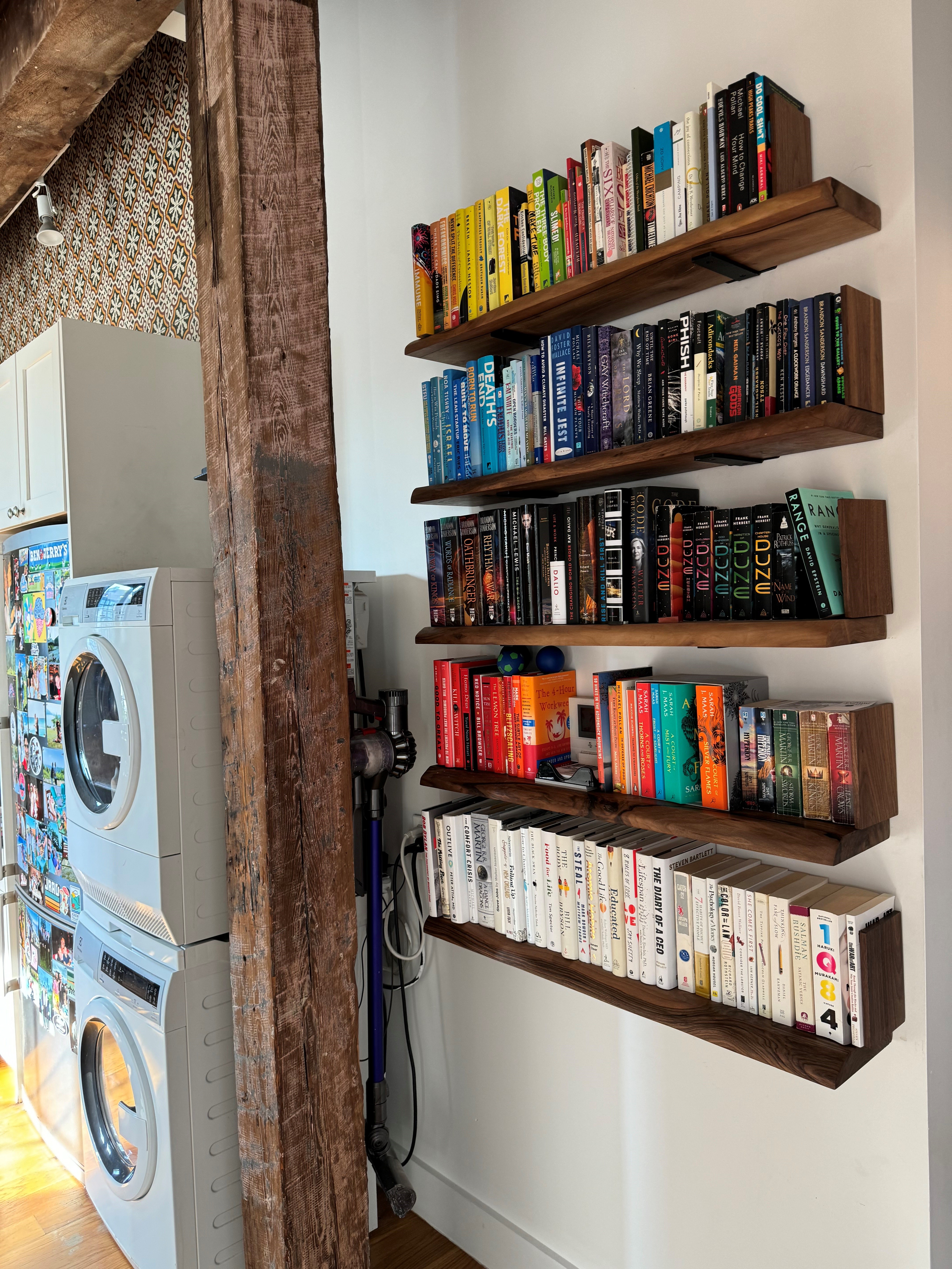 Floating walnut shelves of color-sorted books beside an in-unit stacked washer and dryer