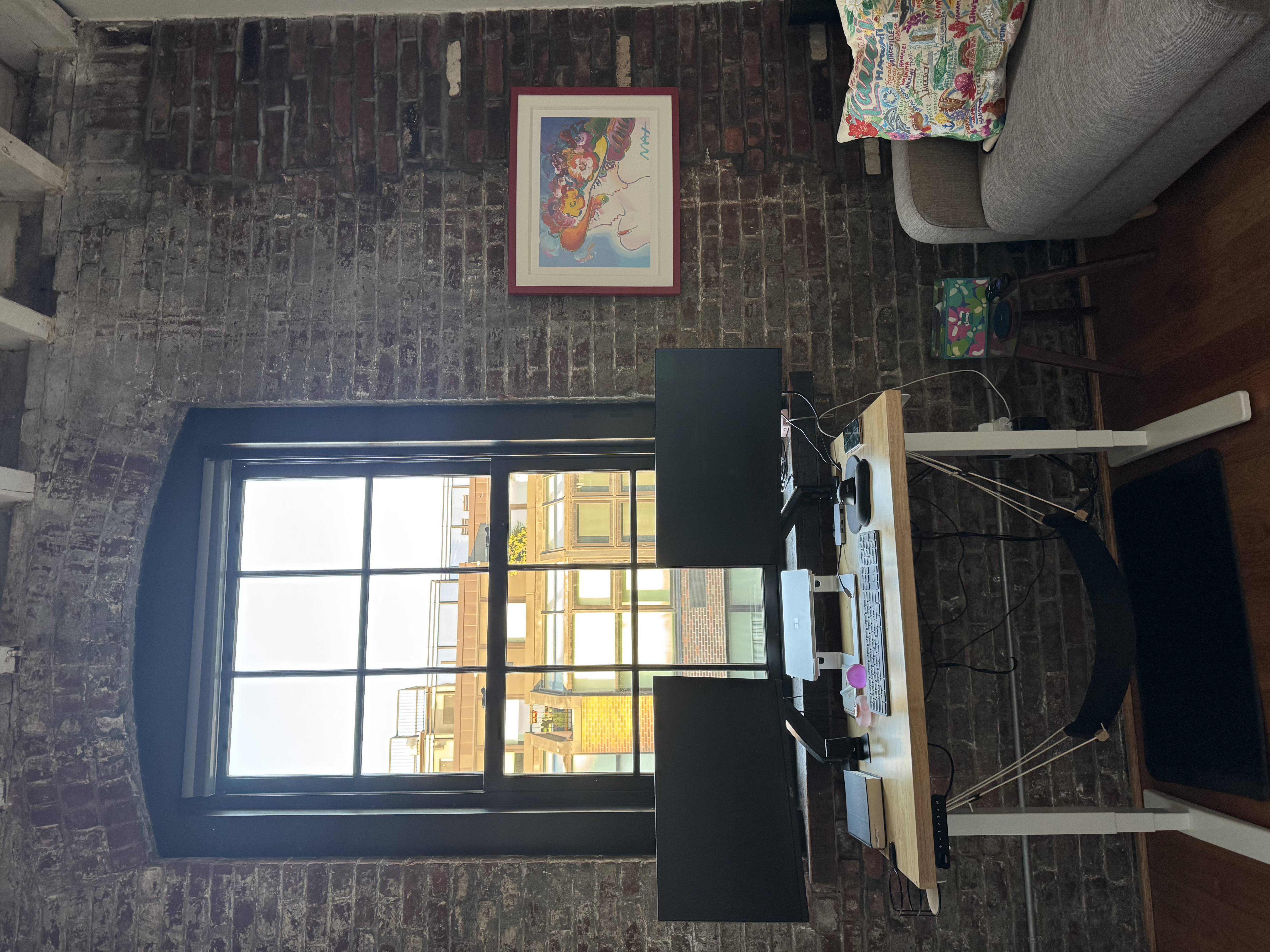 Standing desk with dual monitors under the arched factory window, with a Peter Max print on the exposed brick
