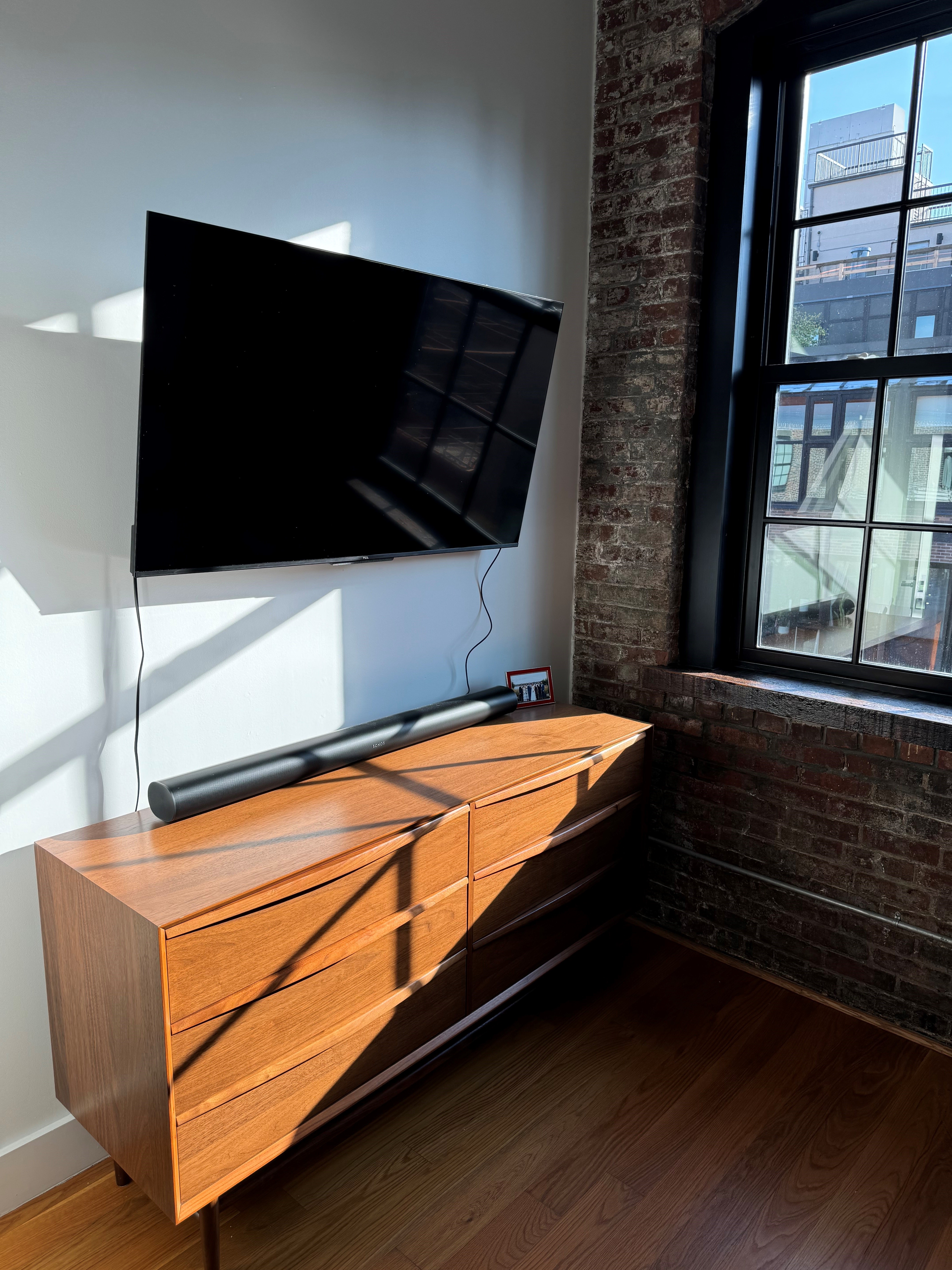 Wall-mounted TV with soundbar on a mid-century dresser beside a brick-framed window
