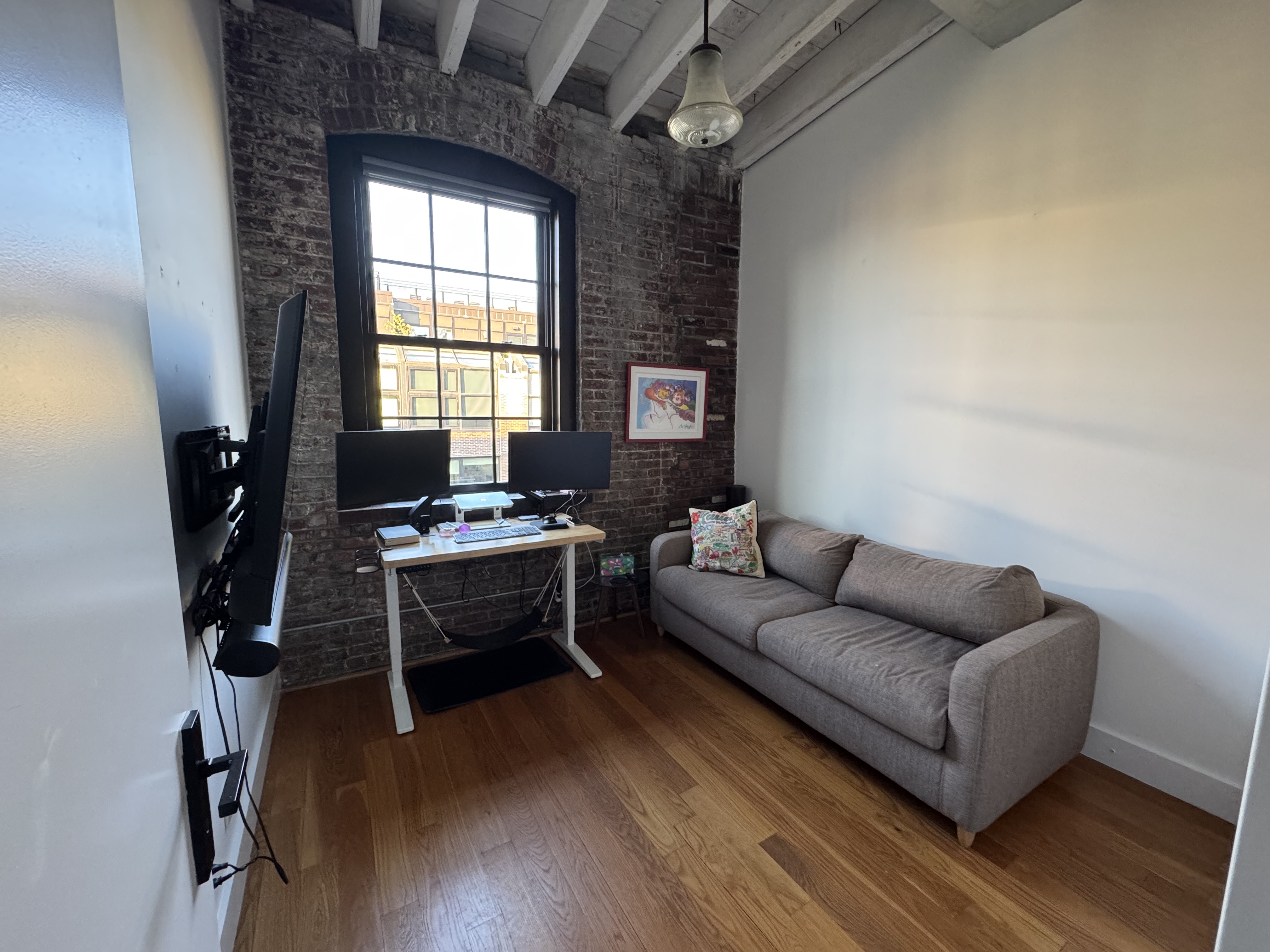 Second bedroom with a wall-mounted TV, standing desk with dual monitors under the arched window, and a pull-out queen couch