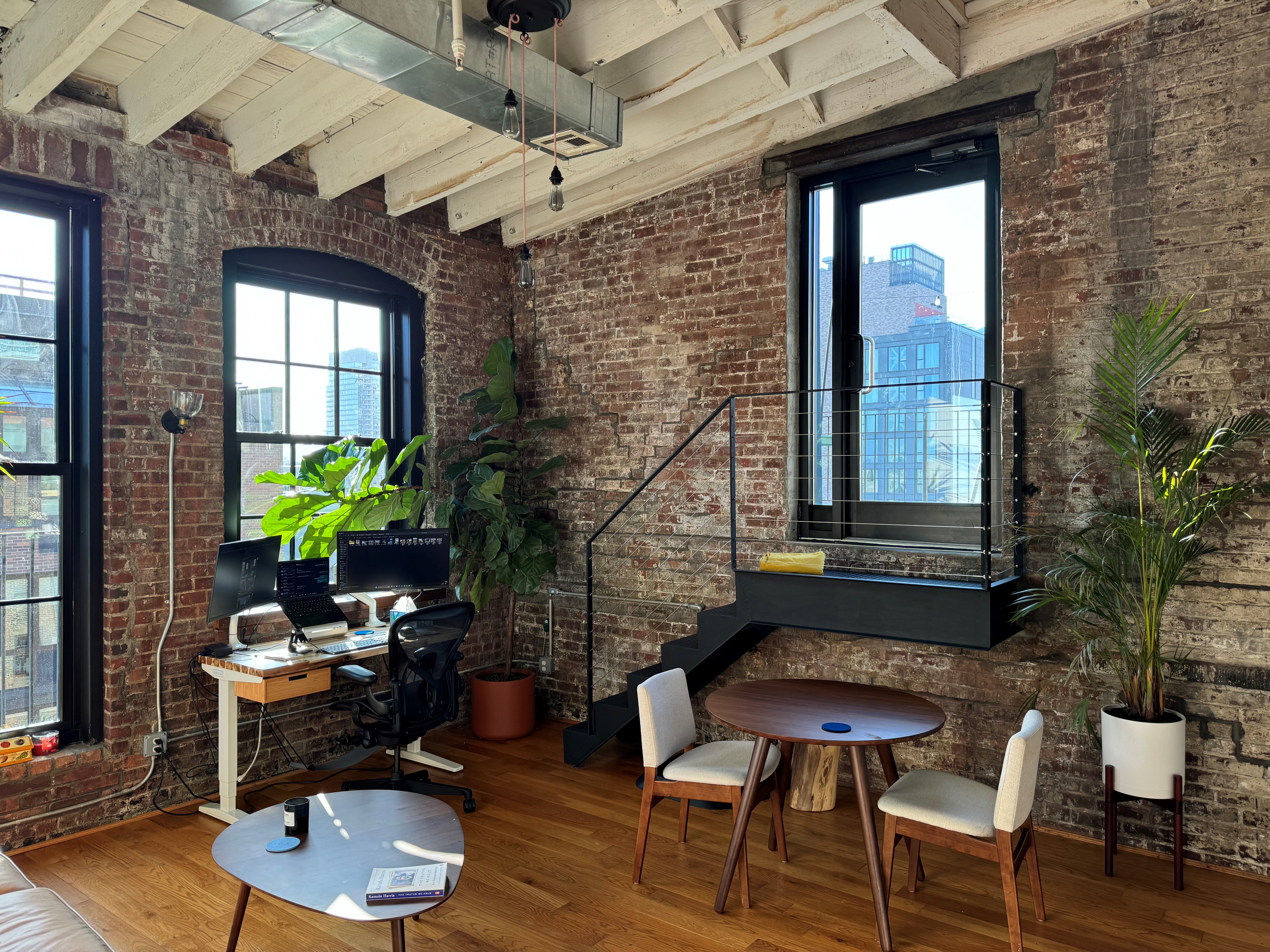 Loft living space with exposed brick walls, arched factory windows, a floating black steel staircase, and plants throughout